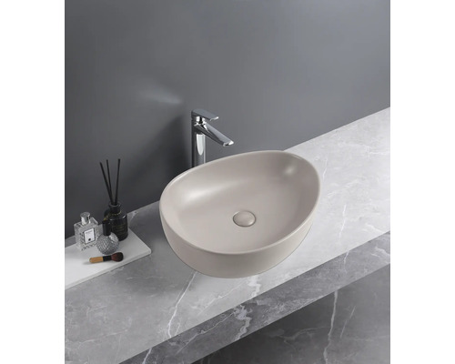 Vasque à poser ovale beige mat sur un plan de travail aspect marbre avec robinetterie chromée.