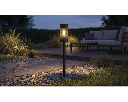 Lampe solaire de jardin à piquer avec ampoule LED et motif en treillis éclairant un chemin de jardin la nuit près d'un salon de jardin.
