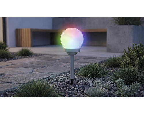 Lampe solaire de jardin en forme de boule avec éclairage RGB multicolore, sur un piquet dans un parterre de gravier avec des plantes.