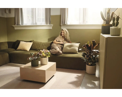 Salon lumineux avec canapé vert olive, femme tenant une tasse, table basse beige et diverses plantes d'intérieur dans un style naturel.