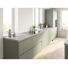 Cuisine moderne avec meubles vert mat sans poignées, plan de travail aspect marbre, plaque de cuisson intégrée et évier sous lumière naturelle.