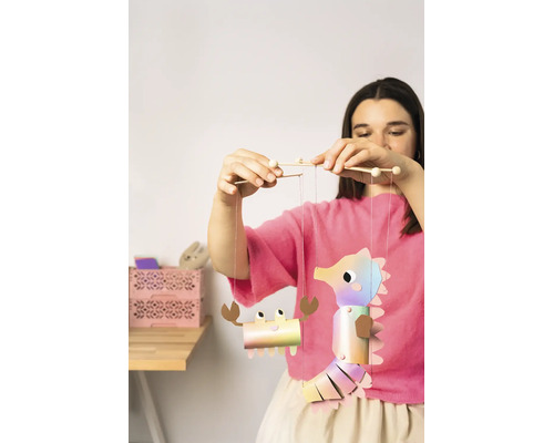 Une femme tient un mobile de bricolage en papier avec un hippocampe et un crabe attachés à des bâtonnets en bois.