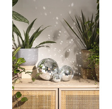 Trois boules à facettes de tailles différentes sur un buffet en bois et rotin, entourées de plantes d'intérieur et de reflets lumineux sur le mur.
