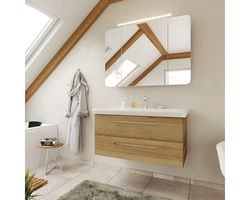 Meuble de salle de bains moderne aspect chêne avec lavabo blanc, armoire de toilette à trois portes et éclairage sous un toit en pente.