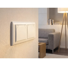 Interrupteur double sur un mur dans un salon lumineux avec canapé et lampadaire.