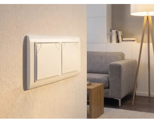 Interrupteur double sur un mur dans un salon lumineux avec canapé et lampadaire.