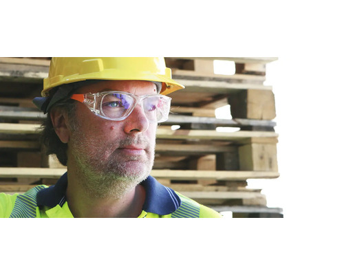 Ouvrier avec casque de chantier jaune et lunettes de protection devant des palettes en bois empilées.