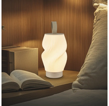 Tragbare LED-Leuchte mit Plissee-Design und Textilschlaufe auf einem Nachttisch aus Holz neben Büchern und Kissen. Warm beleuchtet.