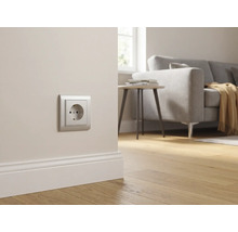 Silberne Schutzkontakt-Steckdose an einer hellen Wand über einer weißen Fußleiste in einem hellen, modern eingerichteten Wohnzimmer.
