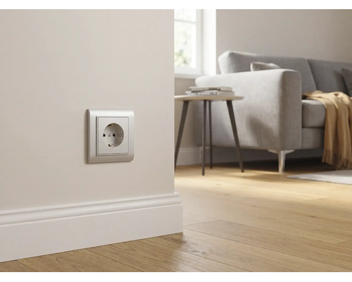 Silberne Schutzkontakt-Steckdose an einer hellen Wand über einer weißen Fußleiste in einem hellen, modern eingerichteten Wohnzimmer.