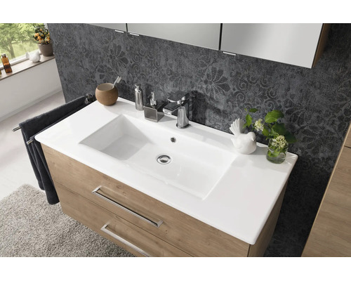 Meuble de lavabo moderne avec vasque blanche, robinetterie chromée et meuble bas en aspect bois devant un mur à motifs.