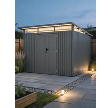 Modernes Gartenhaus aus Metall mit Pultdach, Oberlichtfenstern und Doppeltür auf gepflasterter Fläche in einer hell beleuchteten Gartenanlage.