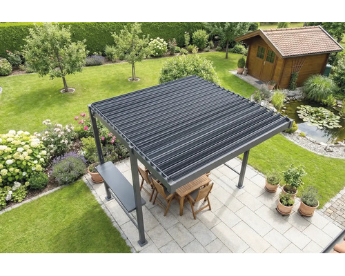 Freistehende Pergola mit Lamellendach auf einer hellen Terrasse im Garten, inklusive Holzmöbeln, angrenzendem Teich und Gartenhaus im Hintergrund.