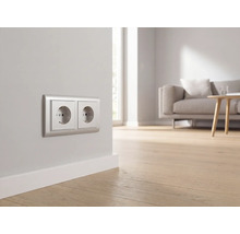Zweifache Wandsteckdose mit silbernem Rahmen in einem hellen, modernen Wohnzimmer mit Holzfußboden und Sofa.