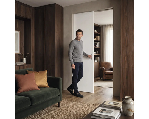 Ein Mann öffnet eine Schiebetür in einem modern eingerichteten Wohnzimmer mit Holzschränken und einem Sofa.