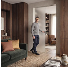 Ein Mann öffnet eine Schiebetür aus satiniertem Glas in einem modernen Wohnzimmer mit dunklen Holzschränken und einem grünen Samtsofa.