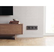 Dreifache Steckdose in Anthrazit an einer hellen Wand in einem modern eingerichteten Wohnzimmer mit Sideboard aus Holz und Parkettboden.