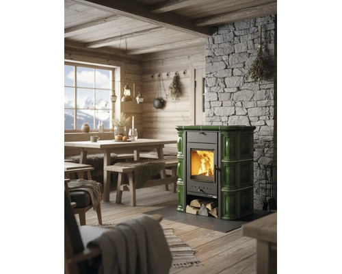 Poêle en faïence verte avec bois brûlant dans une pièce rustique avec mur en pierre et meubles en bois.