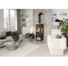 Salon moderne et lumineux avec un poêle à bois noir devant un mur en pierre, canapé blanc, fauteuil, table basse grise et étagères blanches.