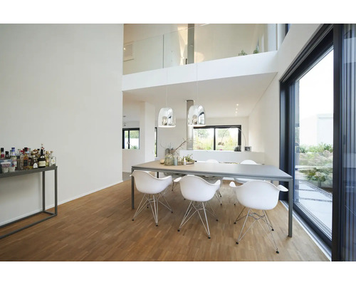 Salle à manger moderne et lumineuse avec une grande table, six chaises blanches, des suspensions argentées, un parquet en bois et une baie vitrée.