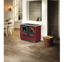 Cuisinière à bois bordeaux avec fenêtre de visualisation et four dans une pièce lumineuse avec parquet et panier de bois de chauffage.