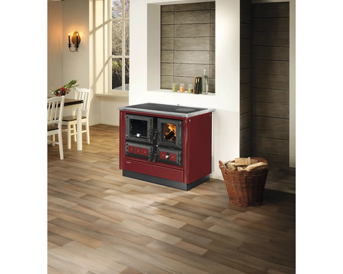 Cuisinière à bois bordeaux avec fenêtre de visualisation et four dans une pièce lumineuse avec parquet et panier de bois de chauffage.