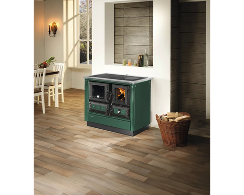 Cuisinière à bois Justus en vert foncé avec feu allumé dans une cuisine lumineuse avec sol en bois et panier de bois de chauffage.