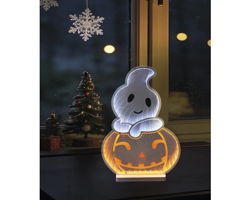 Décoration d'Halloween LED fantôme sur citrouille avec effet miroir infini devant une fenêtre à côté d'un sapin de Noël décoré.