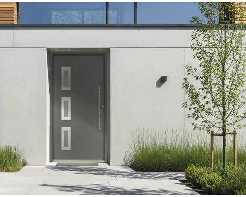 Porte d'entrée moderne anthracite avec trois vitrages et une longue poignée en inox, installée dans une façade claire avec des plantes.