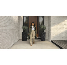 Femme devant une entrée de maison moderne avec des murs en briques blanches, une porte en bois et deux bacs à plantes noirs avec des oliviers.