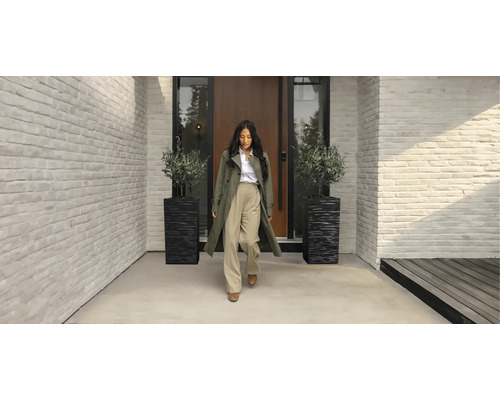 Femme devant une entrée de maison moderne avec des murs en briques blanches, une porte en bois et deux bacs à plantes noirs avec des oliviers.