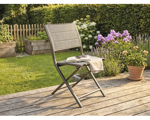 Chaise de jardin pliante avec structure en métal et revêtement en textile sur une terrasse en bois dans un jardin ensoleillé avec des fleurs.