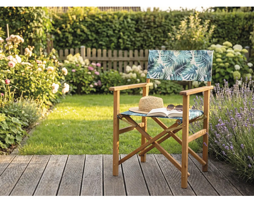 Chaise de metteur en scène en bois avec motif de feuilles tropicales sur une terrasse de jardin avec chapeau de paille et livre.