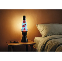 Lampe à lave avec socle noir, liquide bleu et cire rouge sur une table de chevet en bois à côté d'un lit dans une pièce chaleureuse.