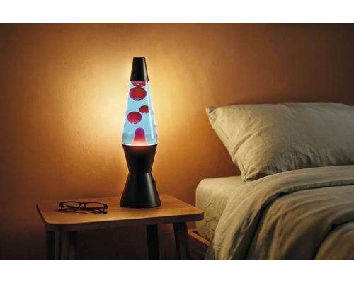 Lampe à lave avec socle noir, liquide bleu et cire rouge sur une table de chevet en bois à côté d'un lit dans une pièce chaleureuse.