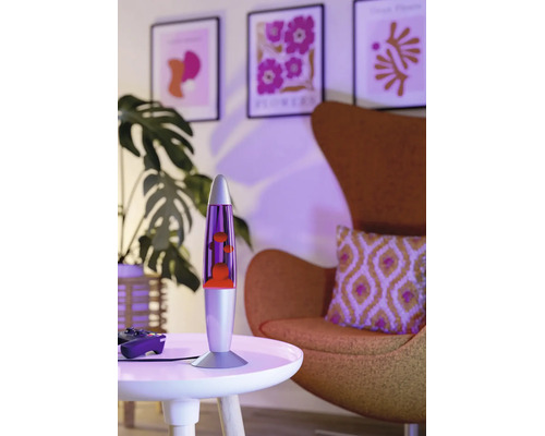 Lampe à lave moderne sur une table ronde dans un salon avec fauteuil, plante et tableaux muraux encadrés dans un éclairage d'ambiance.
