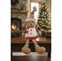 Un ours en peluche vêtu de manière festive assis sur une table dans une pièce chaleureuse avec cheminée et sapin de Noël illuminé.