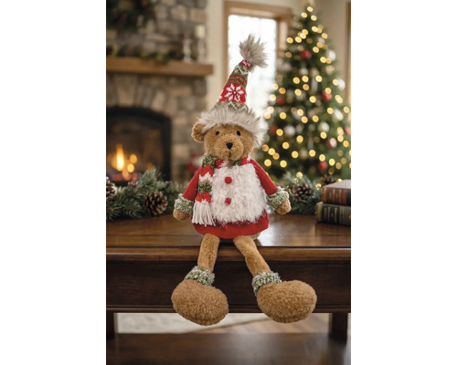 Un ours en peluche vêtu de manière festive assis sur une table dans une pièce chaleureuse avec cheminée et sapin de Noël illuminé.