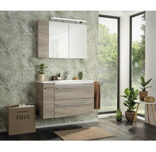 Ensemble de meubles de salle de bains moderne avec meuble sous-vasque aspect bois, lavabo blanc et armoire de toilette devant un papier peint palmier.