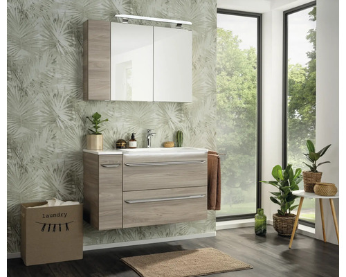 Ensemble de meubles de salle de bains moderne avec meuble sous-vasque aspect bois, lavabo blanc et armoire de toilette devant un papier peint palmier.