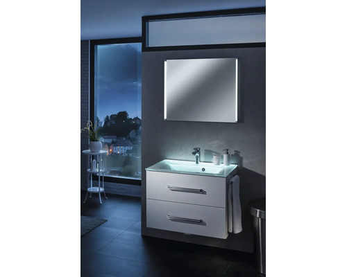 Modern beleuchteter Waschplatz mit weißem Unterschrank, Glaswaschtisch und Spiegel in einem Badezimmer mit Ausblick auf eine nächtliche Stadt.