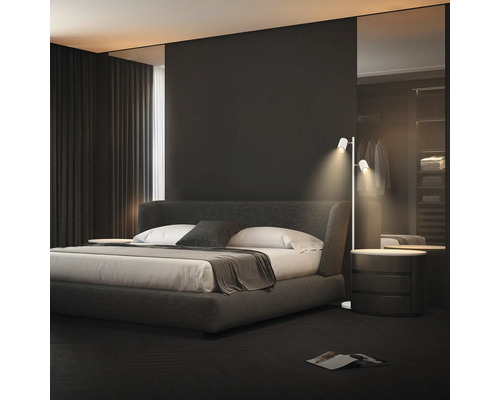Chambre à coucher moderne avec un lit capitonné gris foncé, du linge de lit blanc et un lampadaire blanc fin à deux spots.