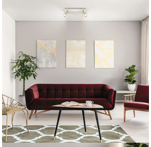 Salon moderne et lumineux avec canapé en velours bordeaux, table basse triangulaire en bois, tapis à motifs, tableaux et plafonnier.