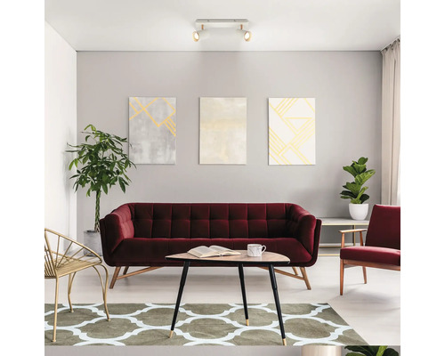 Salon moderne et lumineux avec canapé en velours bordeaux, table basse triangulaire en bois, tapis à motifs, tableaux et plafonnier.