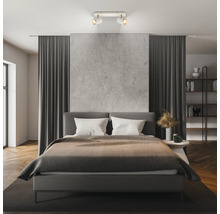 Chambre à coucher moderne avec un spot de plafond à deux lampes éclairant un lit rembourré gris et un mur aspect béton.