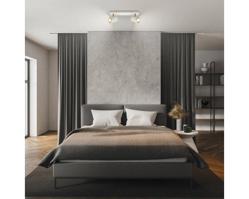 Chambre à coucher moderne avec un spot de plafond à deux lampes éclairant un lit rembourré gris et un mur aspect béton.
