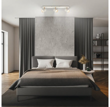 Chambre à coucher moderne avec un spot de plafond à trois lampes, un lit rembourré, un mur aspect béton et des rideaux gris foncé.