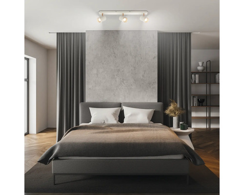 Chambre à coucher moderne avec un spot de plafond à trois lampes, un lit rembourré, un mur aspect béton et des rideaux gris foncé.