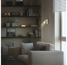 Salon moderne avec un spot mural éclairant un fauteuil beige avec des coussins, à côté d'une étagère murale avec des livres et des décorations.