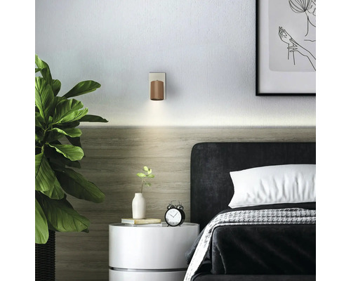 Chambre moderne avec applique murale en bois au-dessus d'une tête de lit sombre, table de chevet blanche avec réveil et grande plante verte.